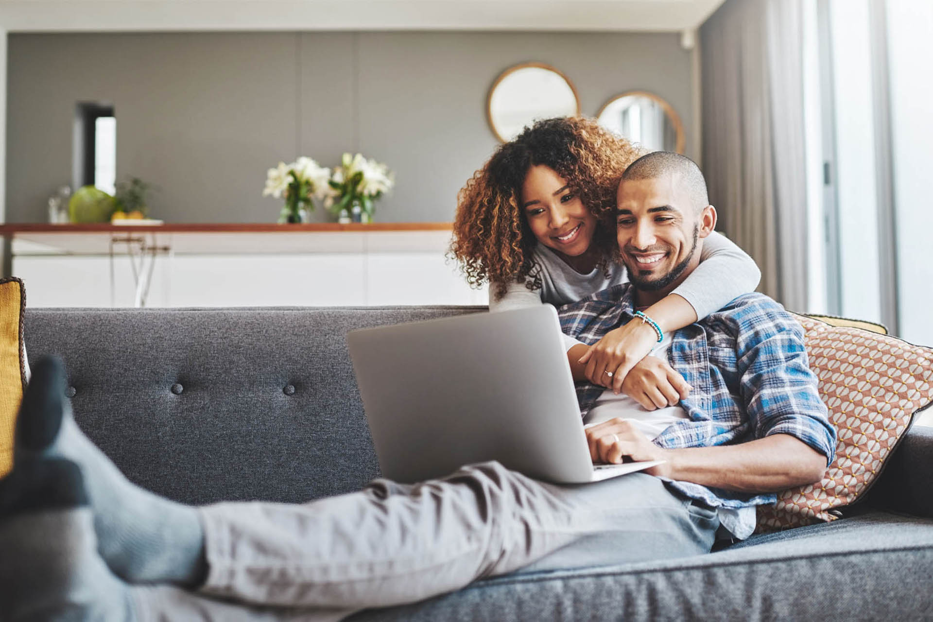 Shot of a young woman hugging her husband while he uses a laptop on the sofa at home